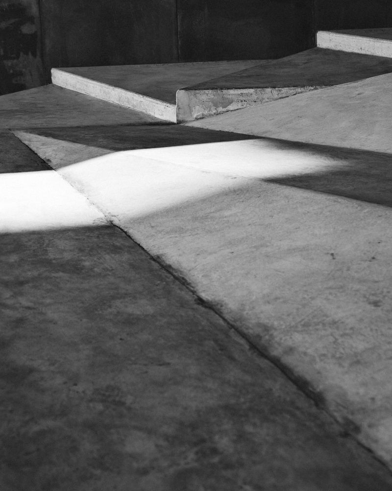 Black and white photograph featuring converging angular concrete steps and a harsh spotlight casting shadows in an indoor setting.