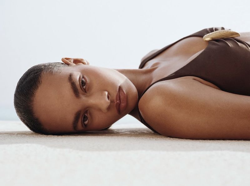 Close-up portrait of a woman lying down with dark hair and a brown swimsuit with a gold ring detail. Soft lighting and skin textures.