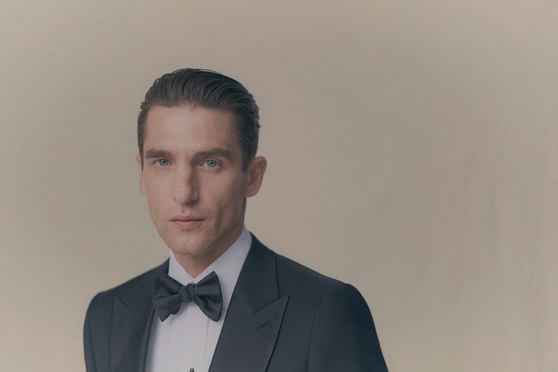 Man wearing a black tuxedo with a white dress shirt and bow tie against a neutral grey backdrop