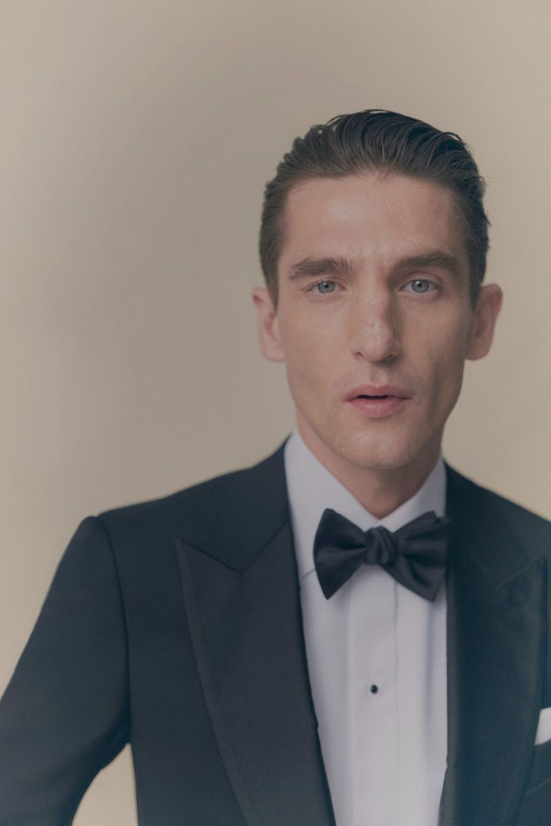 Close-up of adult man in a classic black tuxedo with a bow tie and slicked-back dark hair against a neutral, soft-focus background.