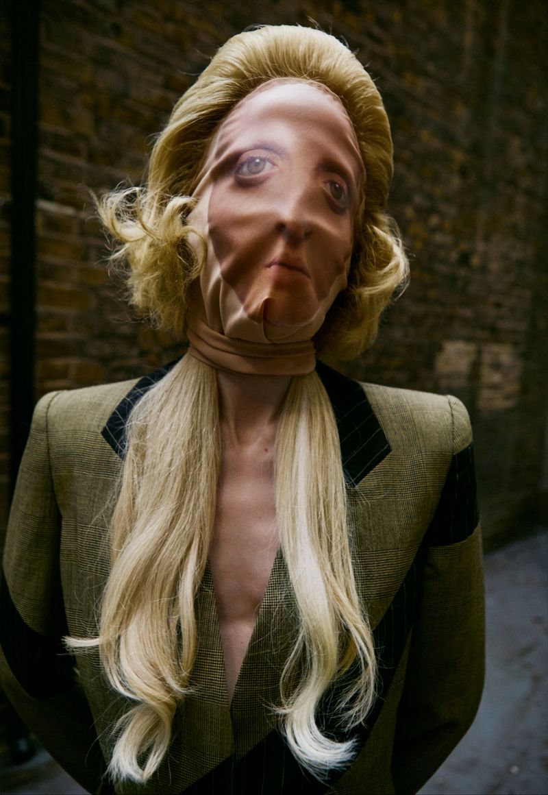 A woman with blonde hair and an olive green blazer is outdoors, with a distorted face under a flesh-colored fabric. Editorial fashion.