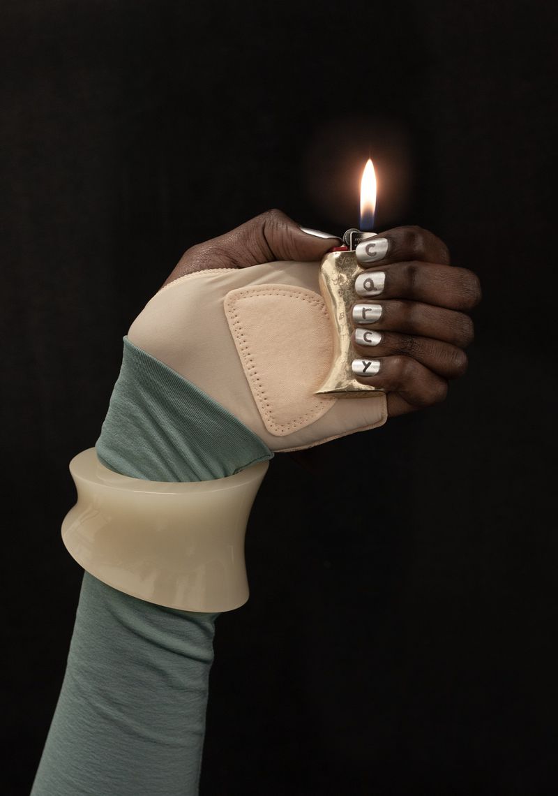 A person with dark skin wearing a glove and a large bangle is holding a lighter with a flame against a solid black background.