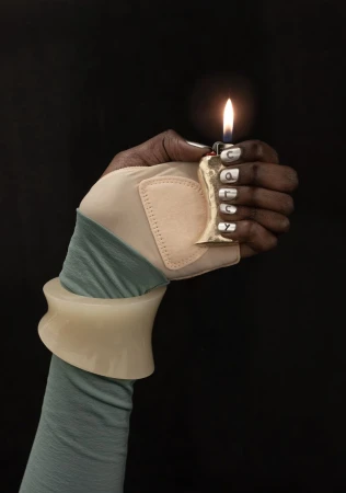 A person with dark skin wearing a glove and a large bangle is holding a lighter with a flame against a solid black background.