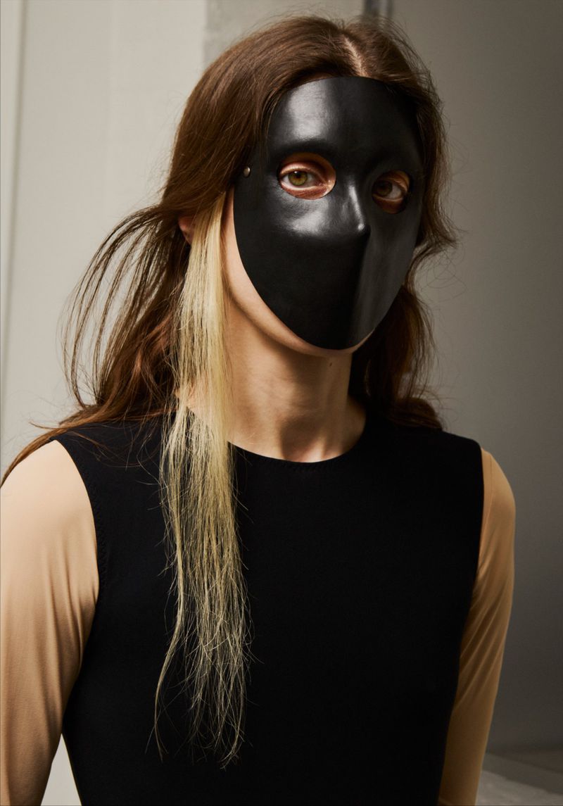 A person with long brown hair is wearing a black mask that covers the nose and mouth, posing indoors against a neutral background.