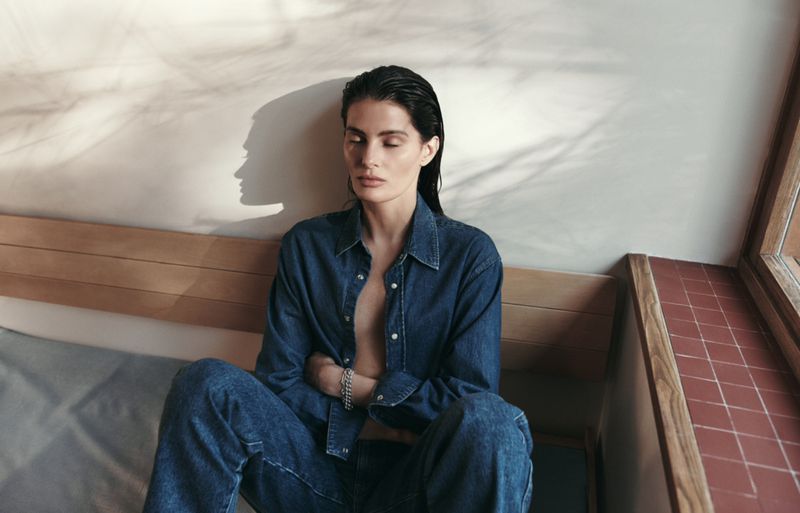 Model Isabeli Fontana wearing a denim shirt and jeans seated against a light wall for Matteau Summer 2026