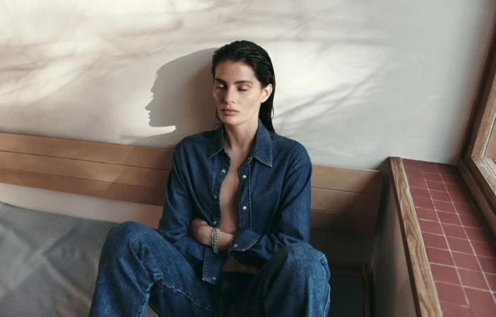 Model Isabeli Fontana wearing a denim shirt and jeans seated against a light wall for Matteau Summer 2026