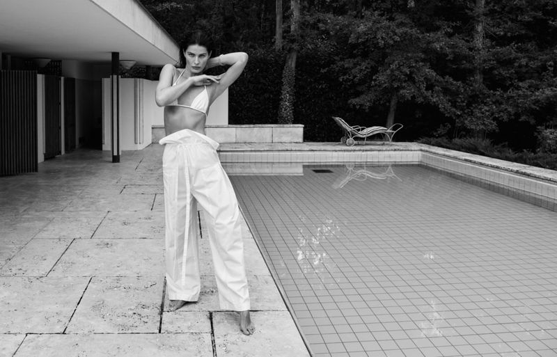 Isabeli Fontana posing by an outdoor swimming pool wearing white trousers and a bikini top for Matteau Summer 2026.