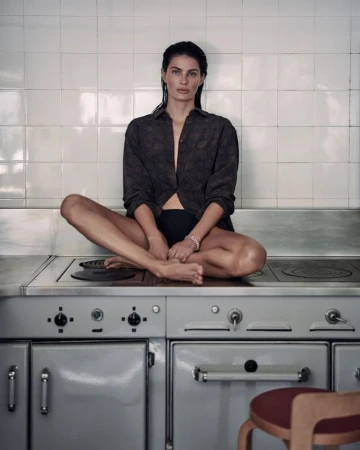Model Isabeli Fontana sitting cross-legged on a kitchen stove wearing a patterned dark shirt and black underwear for Matteau Summer 2026.