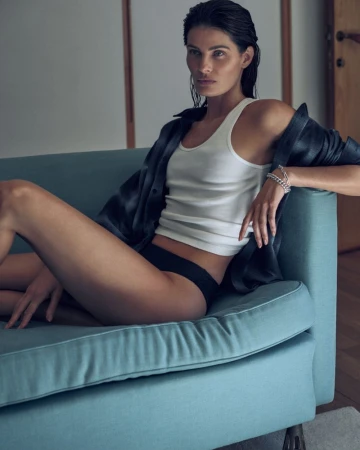 Model Isabeli Fontana sitting on a blue sofa wearing a white tank top and dark shirt for Matteau Summer 2026 campaign.