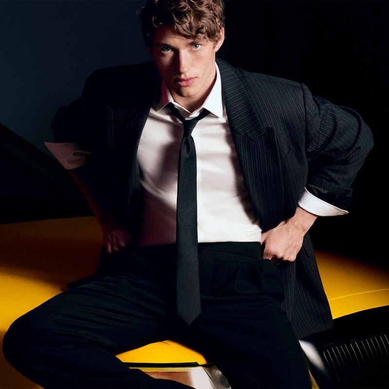 Young man in a black suit jacket and tie leaning on a yellow vehicle against a dark background