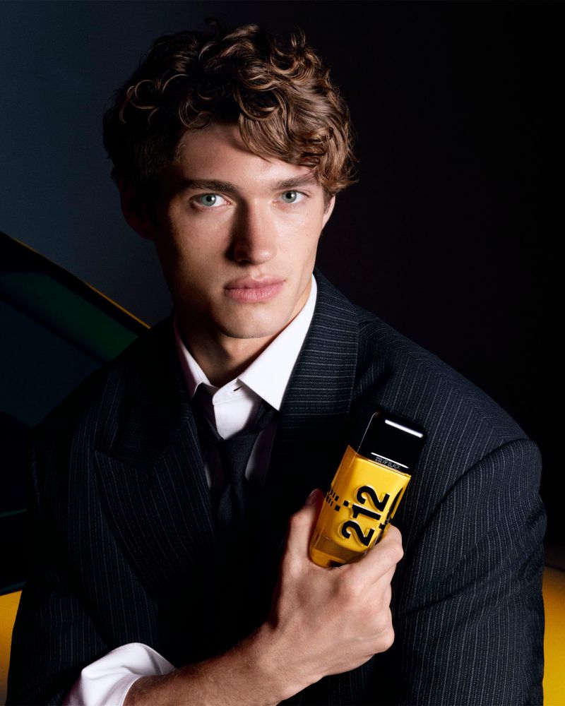 Close-up portrait of a male model in a dark pinstripe suit holding a yellow 212 VIP perfume bottle against a dark background.