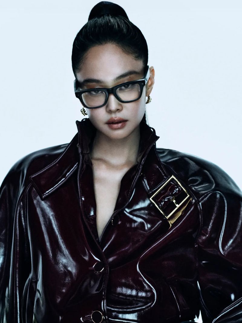 Portrait of a woman with her hair in a bun, wearing black glasses and a shiny burgundy jacket with a gold buckle, against a white background.