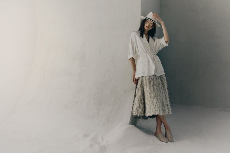 Model wearing a white shirt and textured skirt leaning against a light studio wall