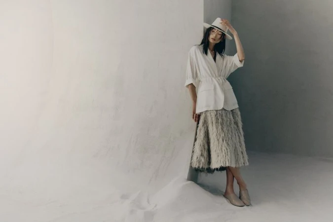 Model wearing a white shirt and textured skirt leaning against a light studio wall