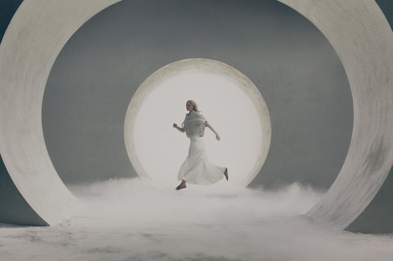 Model in white dress leaping through glowing circular architectural rings framed by low-lying mist