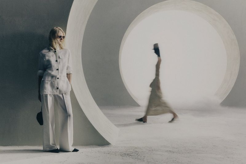 Model in white linen suit standing beside a large circular architectural opening with a blurred figure walking in the background.