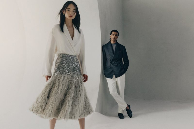 Model in white blouse and sequined feather skirt standing alongside a man leaning against a curved white wall.