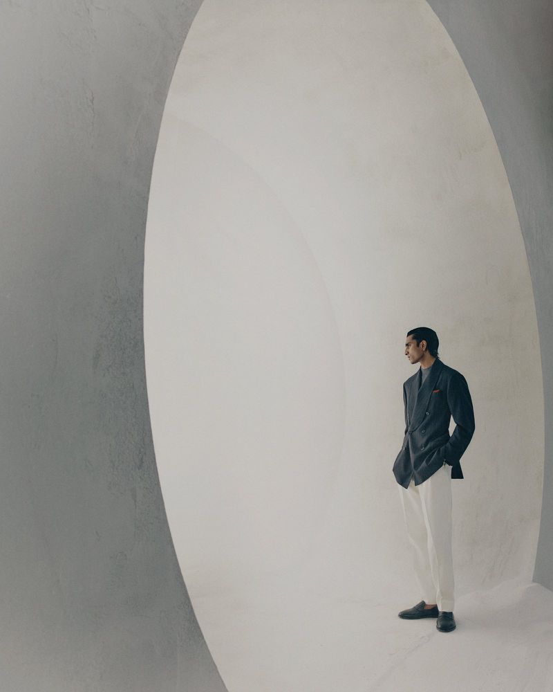 Model standing inside a large architectural white archway wearing a dark blazer and light trousers