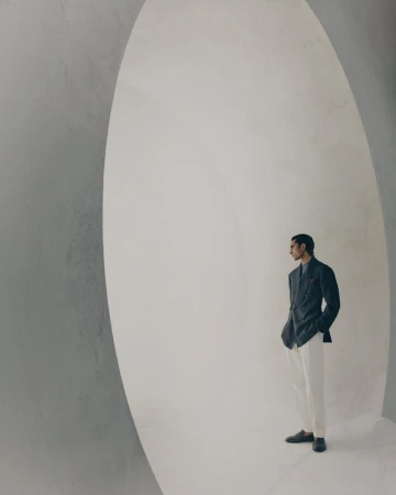 Model standing inside a large architectural white archway wearing a dark blazer and light trousers