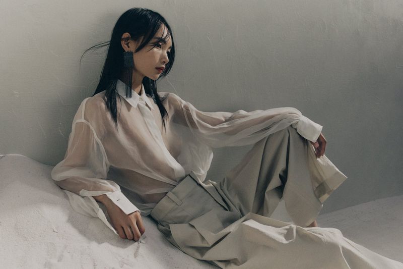 Model in translucent white blouse and pale trousers seated against a textured grey studio wall