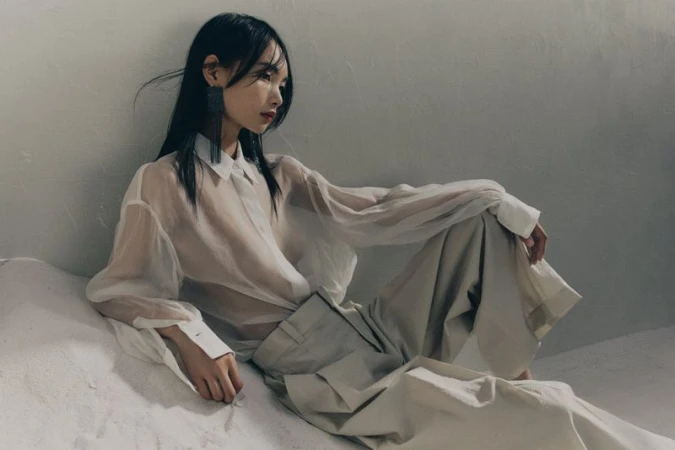 Model in translucent white blouse and pale trousers seated against a textured grey studio wall