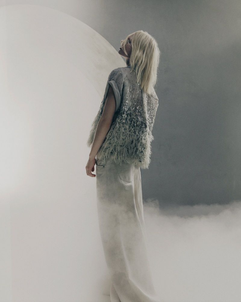 A blonde model wearing a textured grey top and light trousers stands in a misty studio setting, looking upward.