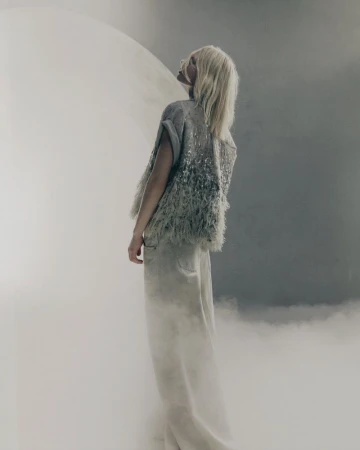 A blonde model wearing a textured grey top and light trousers stands in a misty studio setting, looking upward.