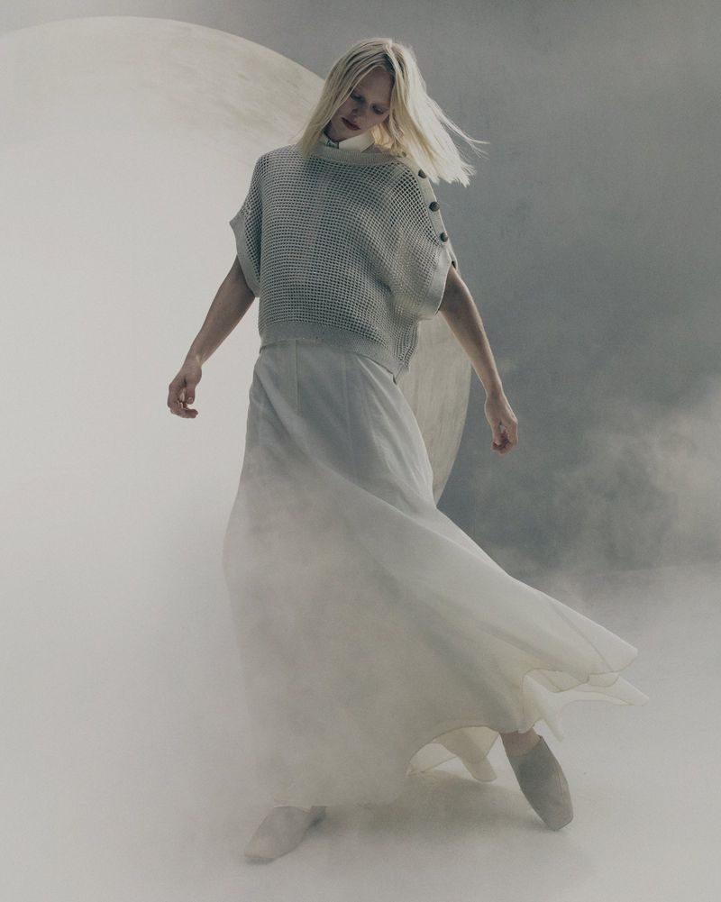 Model in textured grey knit top and long white skirt standing against a circular backdrop with subtle smoke effects.
