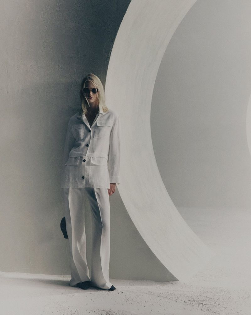 A blonde adult female in a white linen suit and sunglasses stands in front of a large white curved architectural wall in a fashion shot.