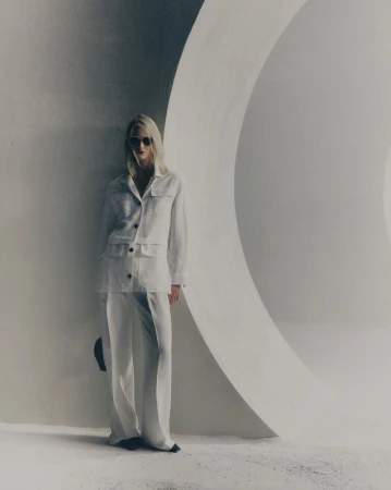 A blonde adult female in a white linen suit and sunglasses stands in front of a large white curved architectural wall in a fashion shot.