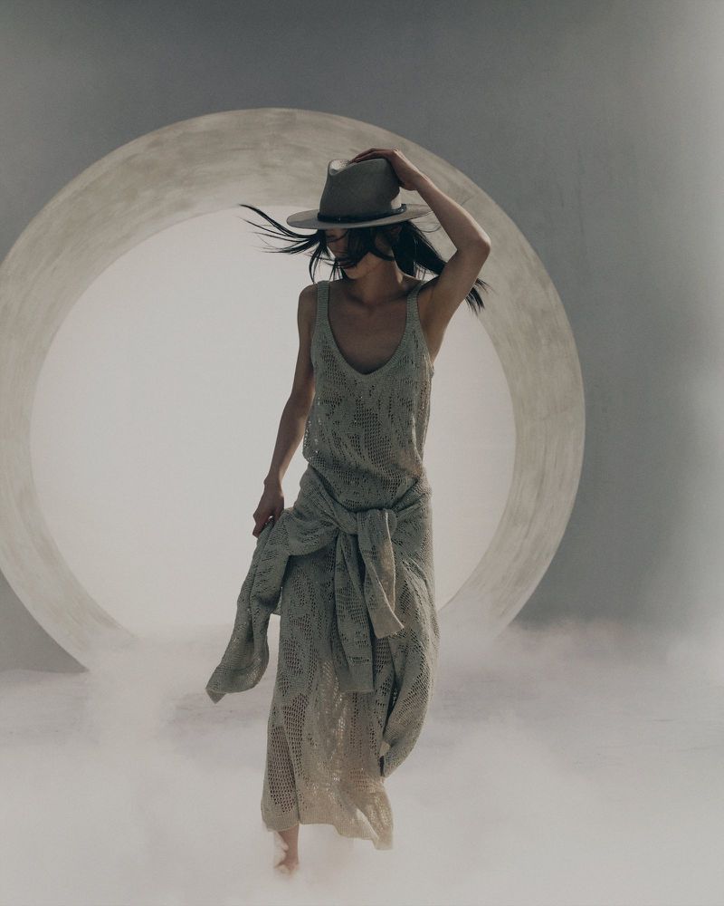 Model in a knit dress and fedora walking through low-lying mist before a large circular studio opening for Brunello Cucinelli.