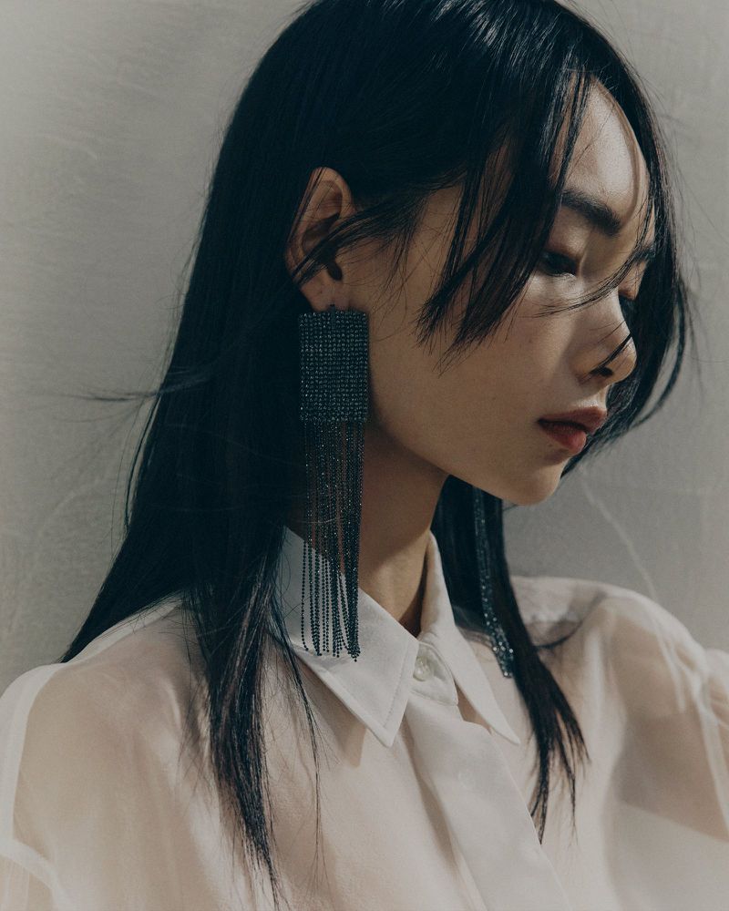 Close-up portrait of an East Asian model wearing long beaded fringe earrings and a white shirt against a neutral textured wall.