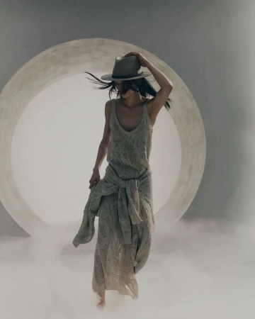 Model in a knit dress and fedora walking through low-lying mist before a large circular studio opening for Brunello Cucinelli.