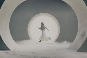Model in white dress leaping through glowing circular architectural rings framed by low-lying mist