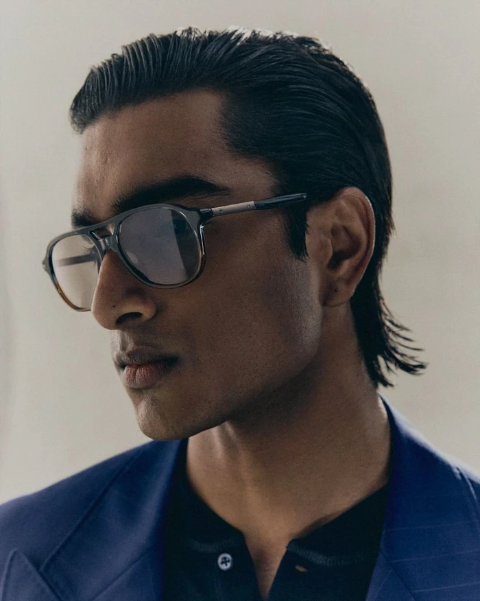 Close-up portrait of a South Asian male model wearing tinted aviator glasses and a blue blazer for Brunello Cucinelli.