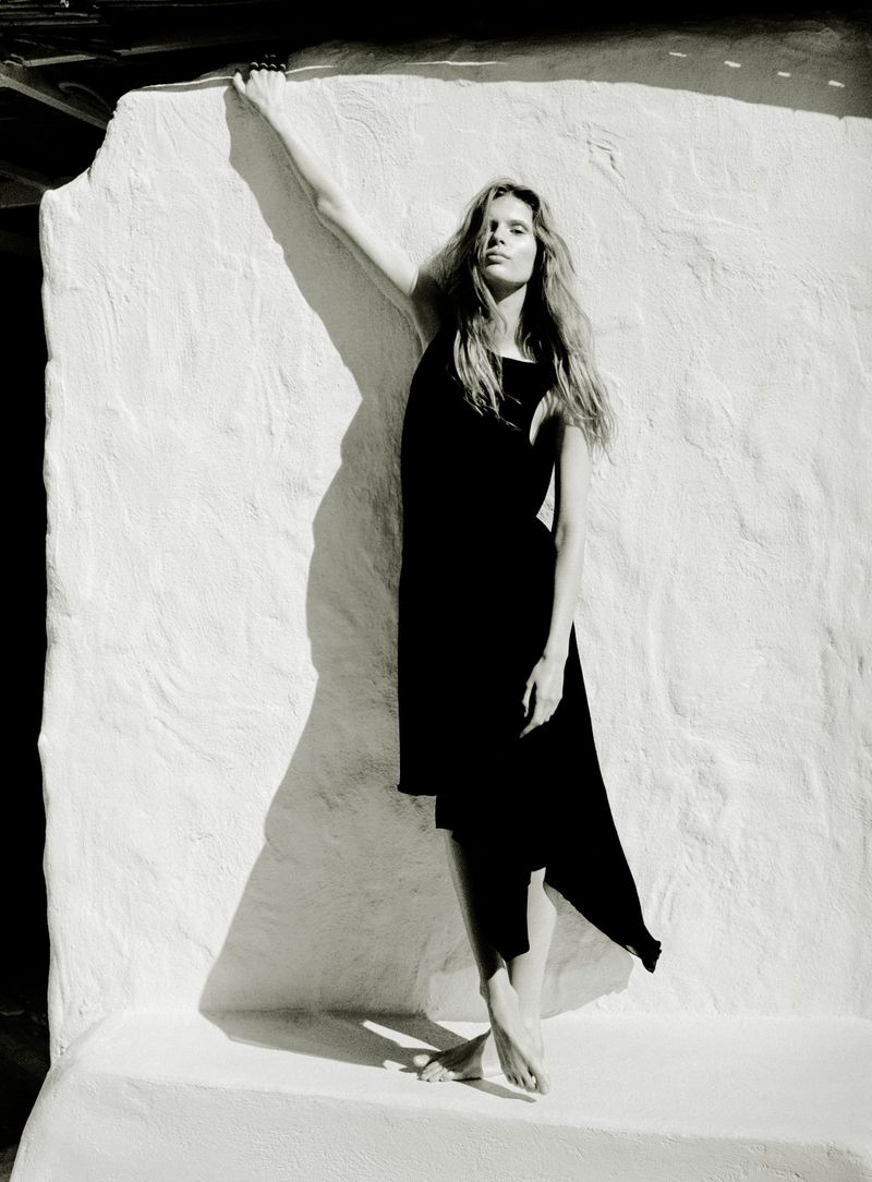 Model in a black dress leaning against a textured white wall outdoors in bright sunlight