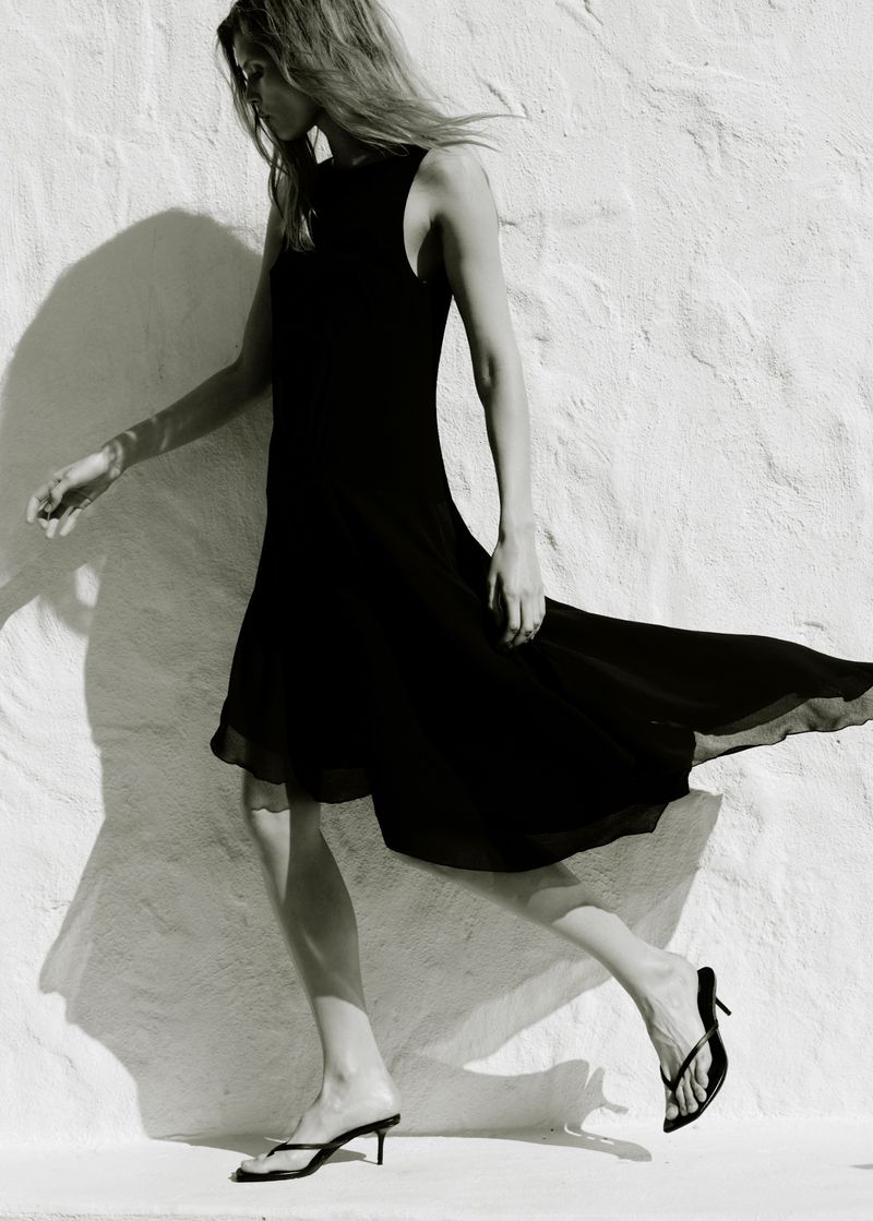 Model wearing a black sleeveless midi dress walking past a textured white wall in bright sunlight