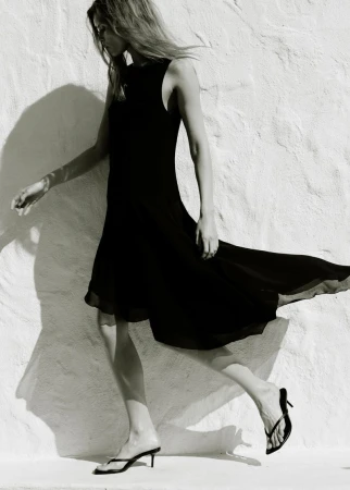 Model wearing a black sleeveless midi dress walking past a textured white wall in bright sunlight