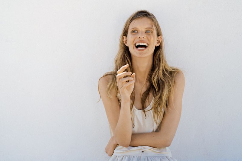 Woman with long blonde hair laughing against a plain white background wearing a light top