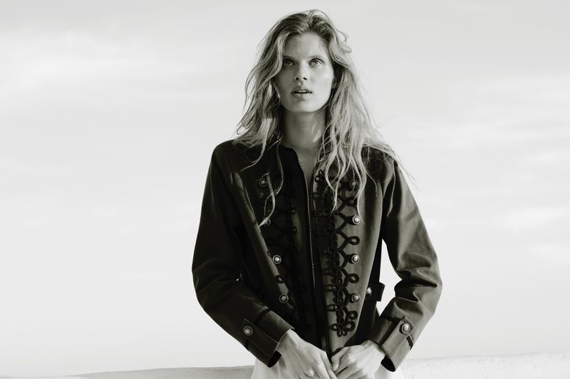 Model wearing a dark military-style jacket with braided closures against an open sky background