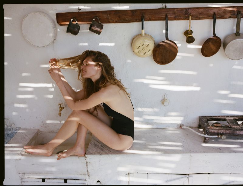 Model Ida Heiner sitting on a rustic kitchen counter wearing a black swimsuit under dappled sunlight for Zara.