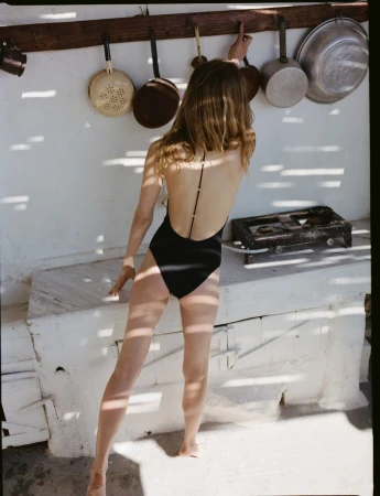 Model wearing a black swimsuit standing outdoors against a white wall with hanging pans and cooking utensils for Zara.