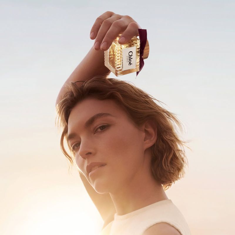 Model holding a Chloé Eau de Parfum Intense bottle overhead against an open sky background
