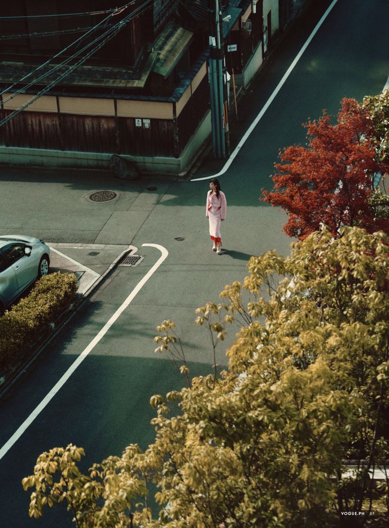 High-angle editorial shot of a woman in pink walking across a quiet city intersection flanked by autumn trees.