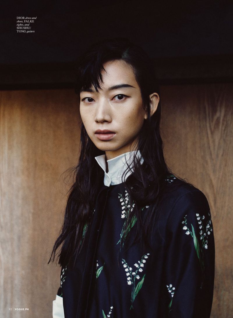 Portrait of a model with dark hair wearing a patterned top with white collar against a dark wooden wall.