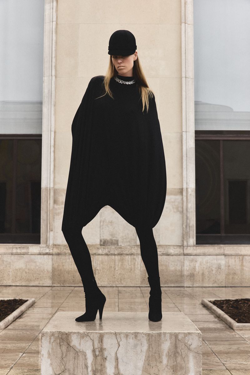Model wearing an oversized black outfit and cap standing on a stone pedestal before building exterior.