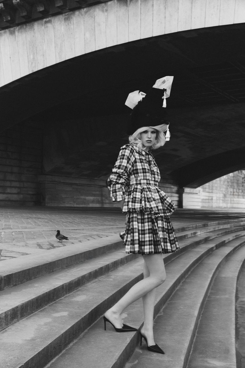 Model in plaid dress and elaborate hat standing on stone steps under a bridge