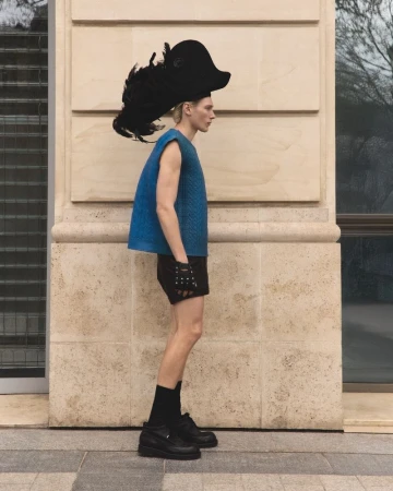 Profile shot of a male model wearing a blue textured top, dark shorts, black gloves, and a large feathered hat against a stone wall.