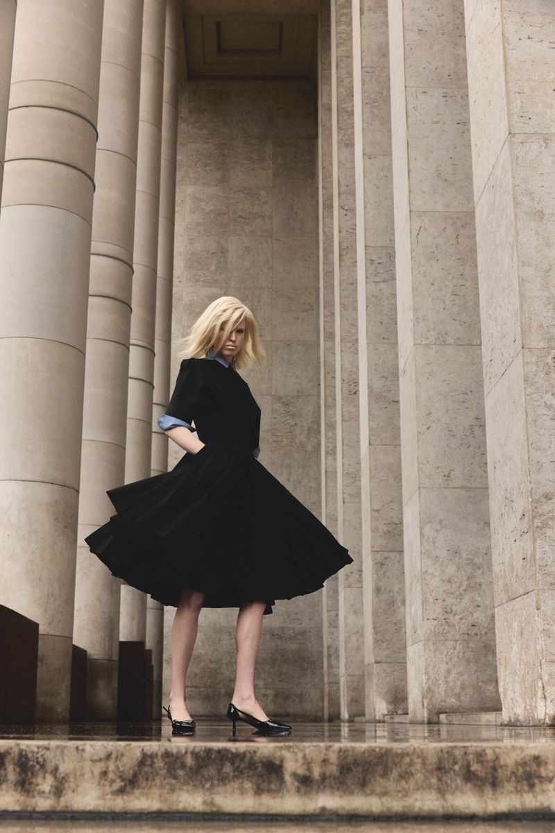 Model in a black flared dress and heels standing amid massive stone columns for Numero Netherlands editorial.