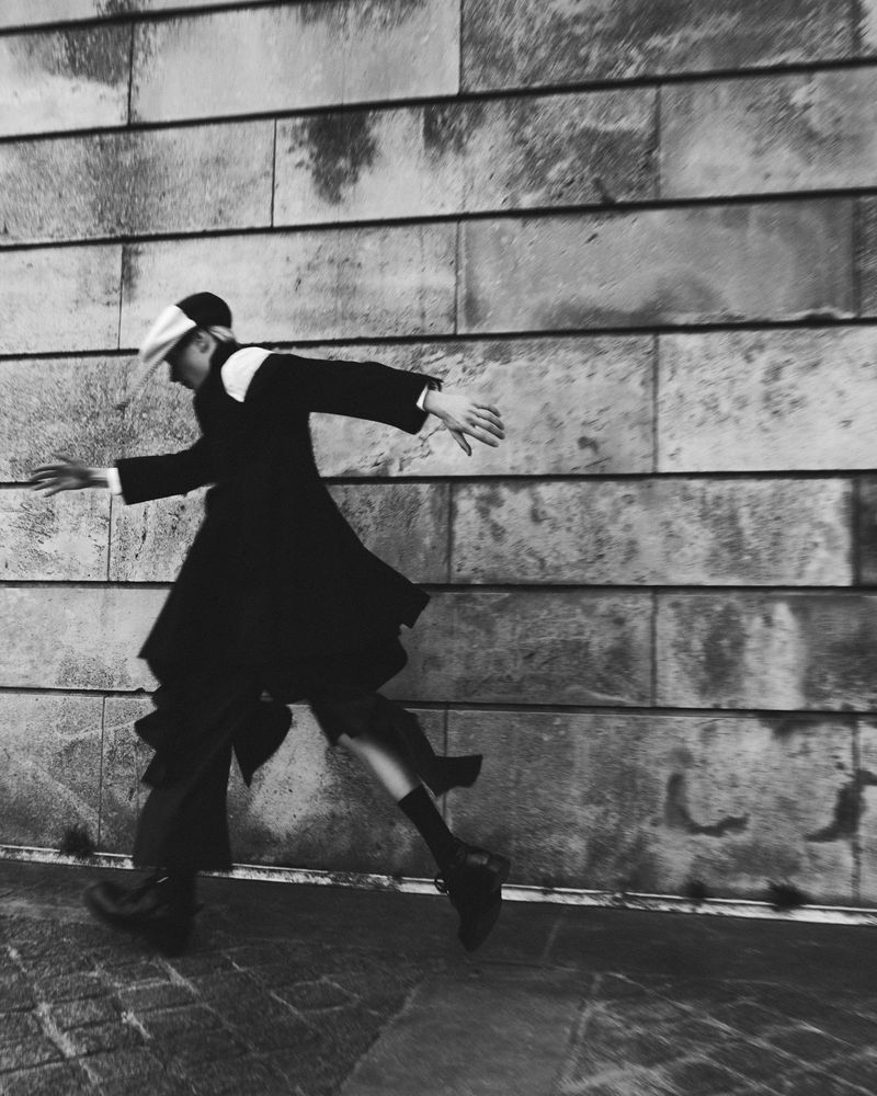 Model in motion wearing a dark coat and cap against a large stone block wall